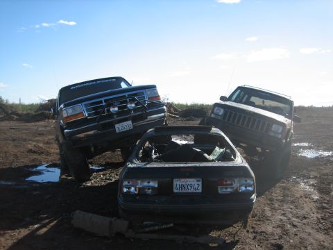 1995 Ford Bronco - Three vehicles, including a black Ford Bronco and a Jeep Cherokee, are positioned on a muddy, outdoor lot under a bright blue sky.