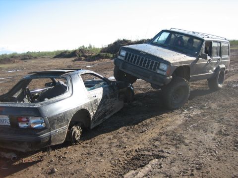 1995 Ford Bronco - A Jeep Cherokee is positioned on top of a heavily damaged Mazda RX-7 in a muddy, outdoor area.