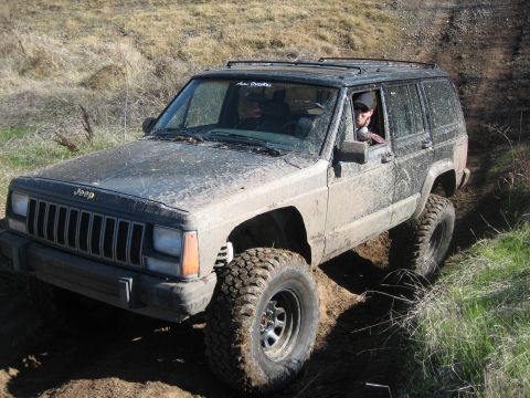 1995 Ford Bronco - A muddy Jeep Cherokee XJ navigates an off-road trail with large tires.