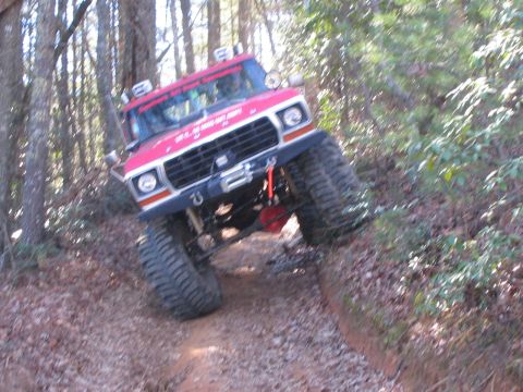 1979 Ford Bronco - A red and white lifted Ford truck with oversized off-road tires and a front winch bumper climbs a rutted dirt trail through a wooded area.…