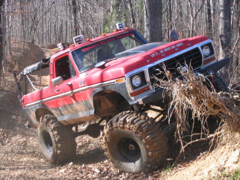 1979 Ford Bronco - Modified red Ford F-Series truck navigating a difficult off-road trail with significant tree roots.