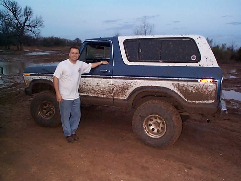 Man in white shirt standing beside a mud-splattered blue and white Ford Bronco