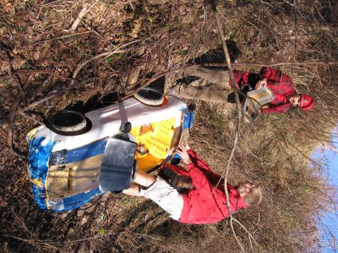 1997 Ford Probe - Two individuals are near an overturned vehicle in a natural setting.