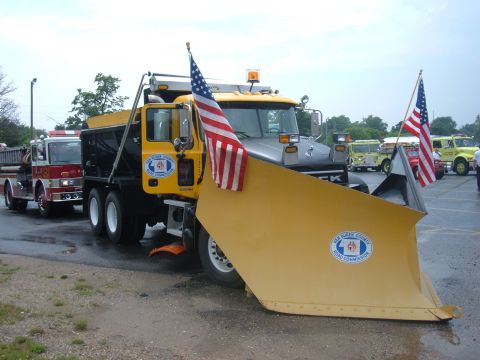 1985 Ford F-150 - A large yellow snow plow truck is adorned with American flags, parked alongside a fire truck and other vehicles.