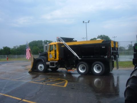1985 Ford F-150 - A large snow plow truck is parked on a wet surface, displaying an American flag on its front plow.