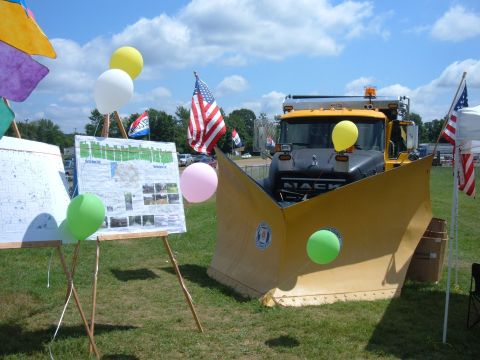 1985 Ford F-150 - A Mack snow plow truck is displayed outdoors with balloons and American flags, alongside informational posters.