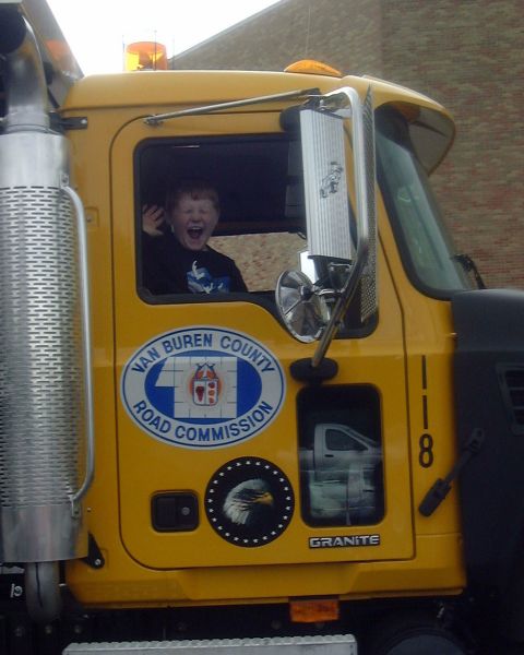 1985 Ford F-150 - A child is excited in the driver's seat of a yellow truck with a road commission logo.