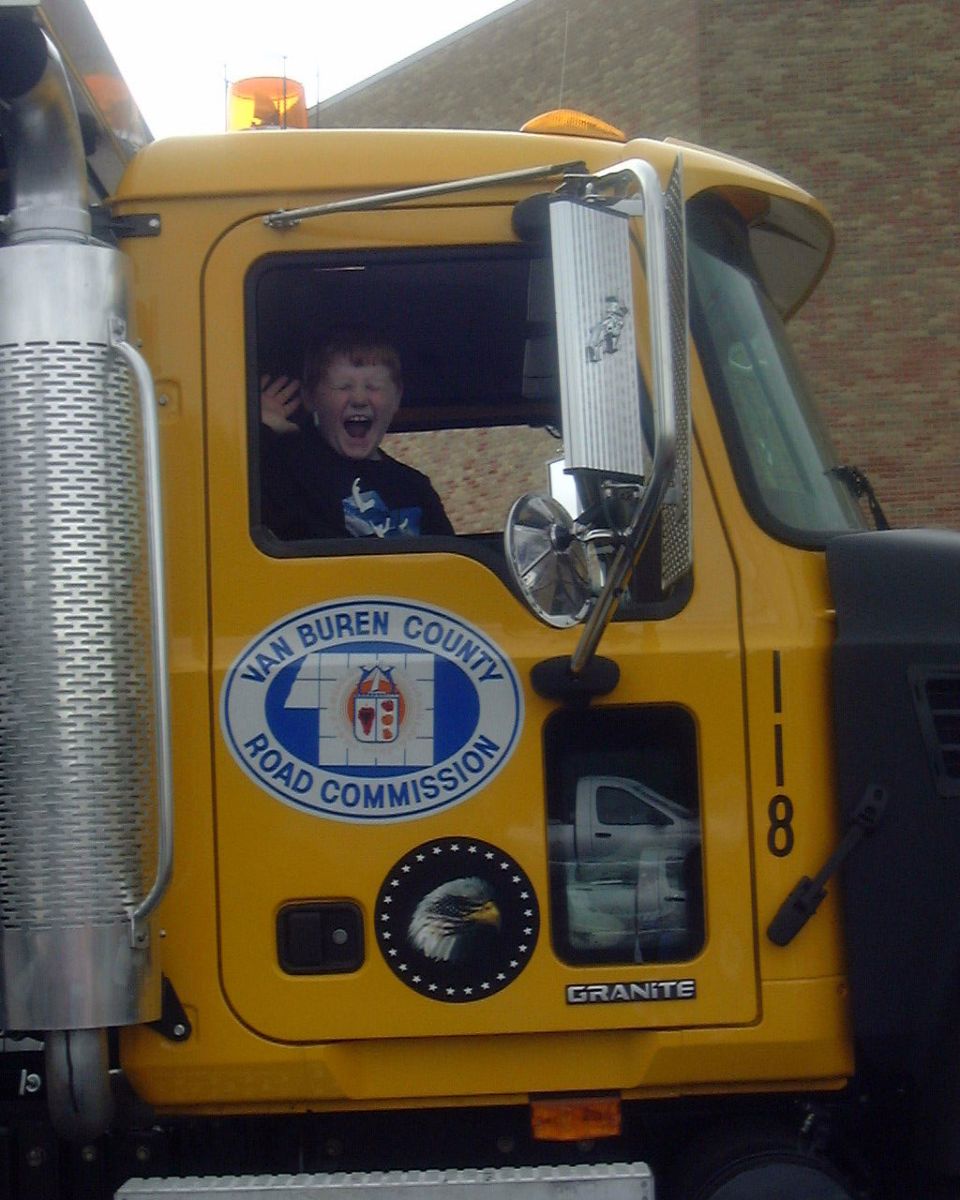Child with mouth open wide in the cab of a yellow truck with 'Van Buren County Road Commission' logo