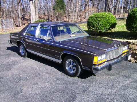 1982 Ford Crown Victoria LTD - A dark brown sedan with a chrome grille and trim sits parked on a paved surface in front of trees.