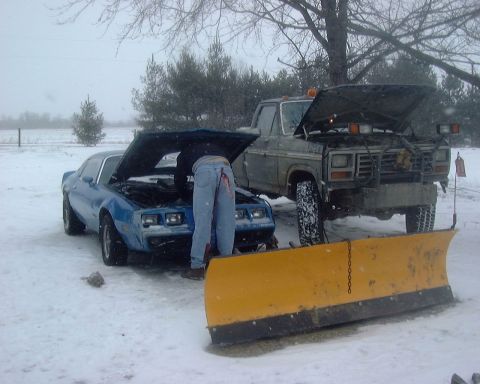 1985 Ford F-150 - A person works on a blue coupe in the snow next to a snow plow truck.