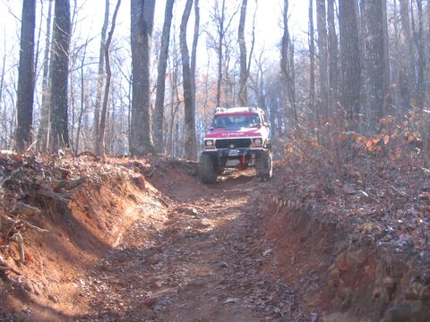 1979 Ford Bronco - A red off-road SUV navigates a steep, muddy trail through a dense forest.