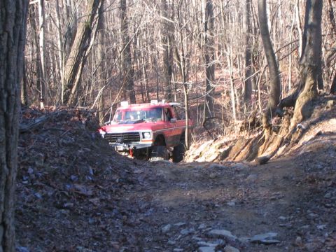 1979 Ford Bronco - Red 1979 Ford Bronco navigates a rough trail in a forest during daylight.