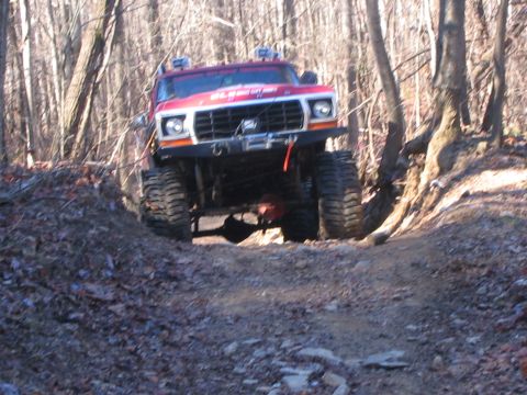 1979 Ford Bronco - A red off-road truck navigates a wooded trail, its large tires gripping the dirt.