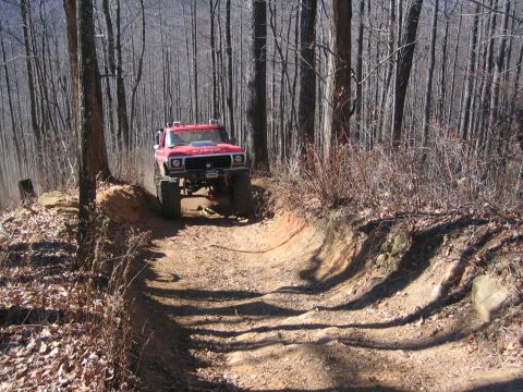 1979 Ford Bronco - A lifted red truck navigates a rutted path through a wooded area.