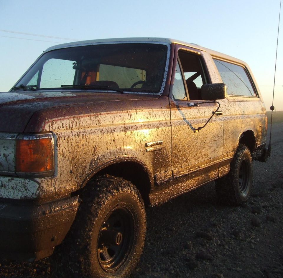 Mud-covered two-door Ford Bronco, brown and white, parked on gravel