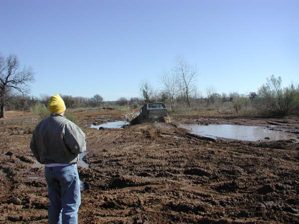 Man in yellow beanie watches a truck cross a distant mud pit in an open field