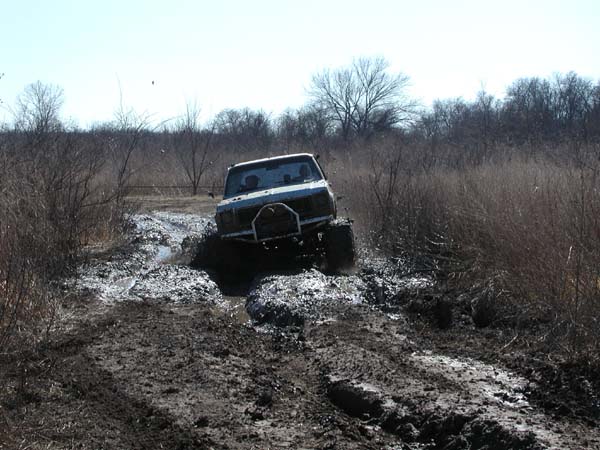 Mud-soaked SUV with brush guard crawling head-on through a rutted trail lined with bare brush