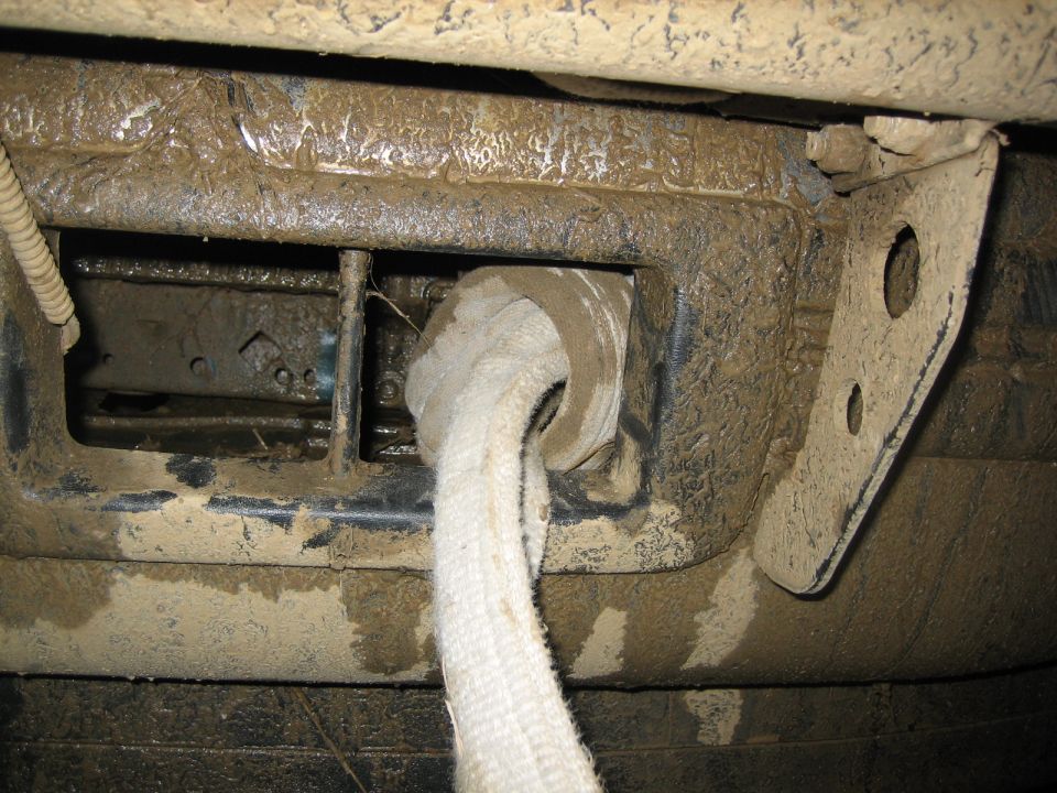 Mud-covered tow hook assembly on a vehicle frame, with a white tow strap looped through it