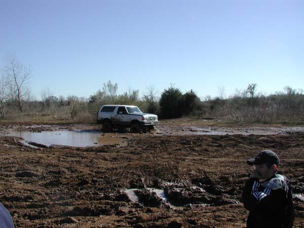 White lifted Ford Bronco parked in muddy clearing, young man in cap watching at right