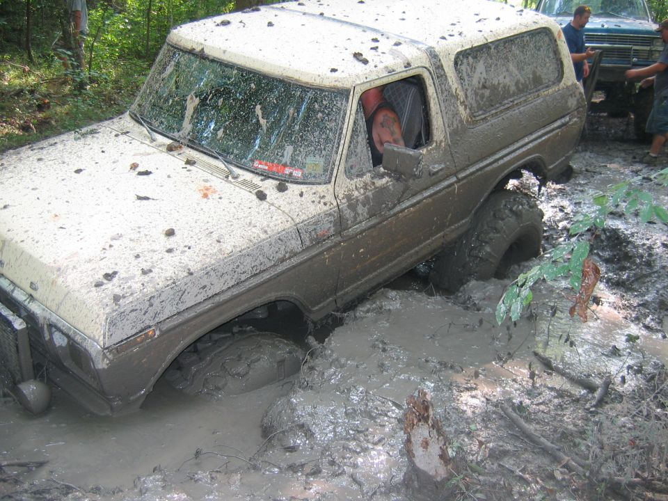 Mud-covered 1978 Ford Bronco stuck in mud with driver visible, another vehicle behind