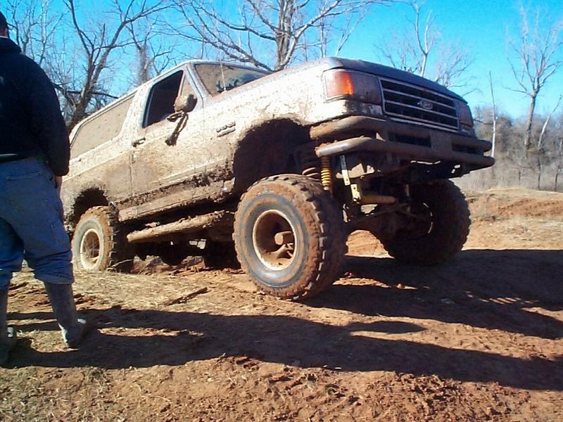 Heavily lifted muddy Ford Bronco on big tires flexing over a dirt mound, bystander at left