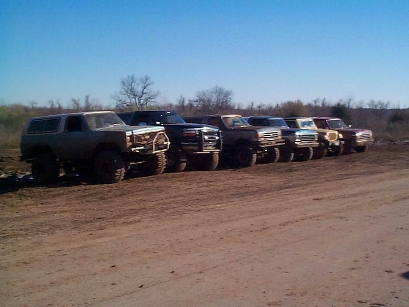 Row of lifted trucks and a Bronco parked nose-out on dirt under clear blue sky