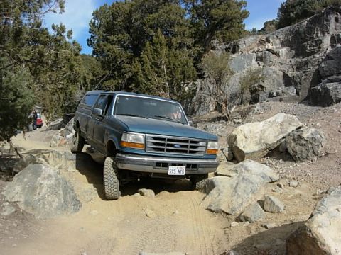 1987 Ford Bronco - A blue SUV with a camper shell is shown off-roading on a rocky terrain, with another vehicle visible in the background.