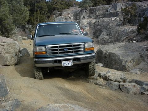 1987 Ford Bronco - A blue Ford F-Series truck navigates a challenging rocky terrain on an off-road trail.