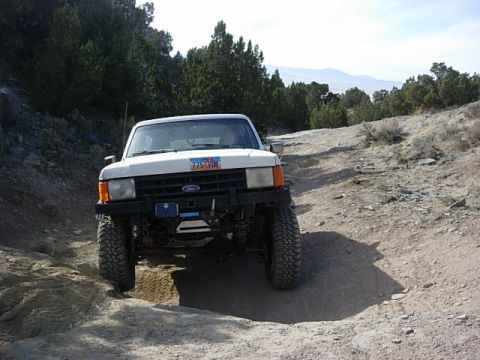 1987 Ford Bronco - A white 1987 Ford Bronco navigates a rocky off-road trail under a clear sky.