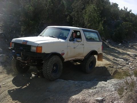 1987 Ford Bronco - A white 1987 Ford Bronco navigates a , off-road trail with large tires and a lifted suspension.