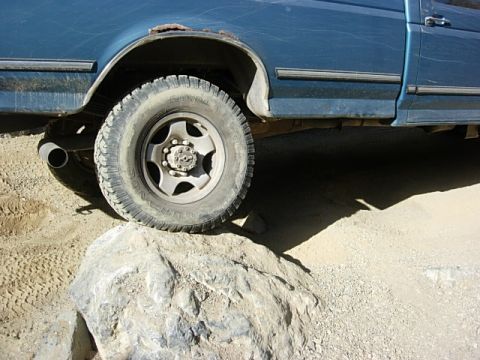 1987 Ford Bronco - Close-up of a blue pickup truck's wheel on a rock, with visible rust on the fender.