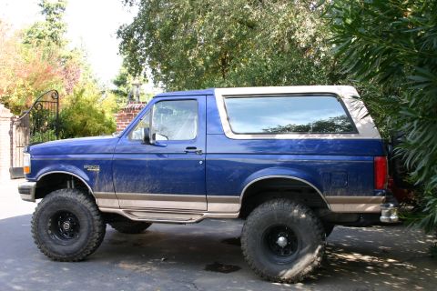 1996 Ford Bronco - A blue Ford Bronco with a tan stripe and removable top is parked on a paved surface.