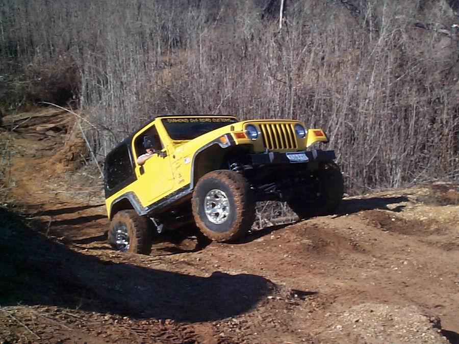 Yellow Jeep Wrangler with Diamond Off Road decal climbing a dirt ledge in the woods