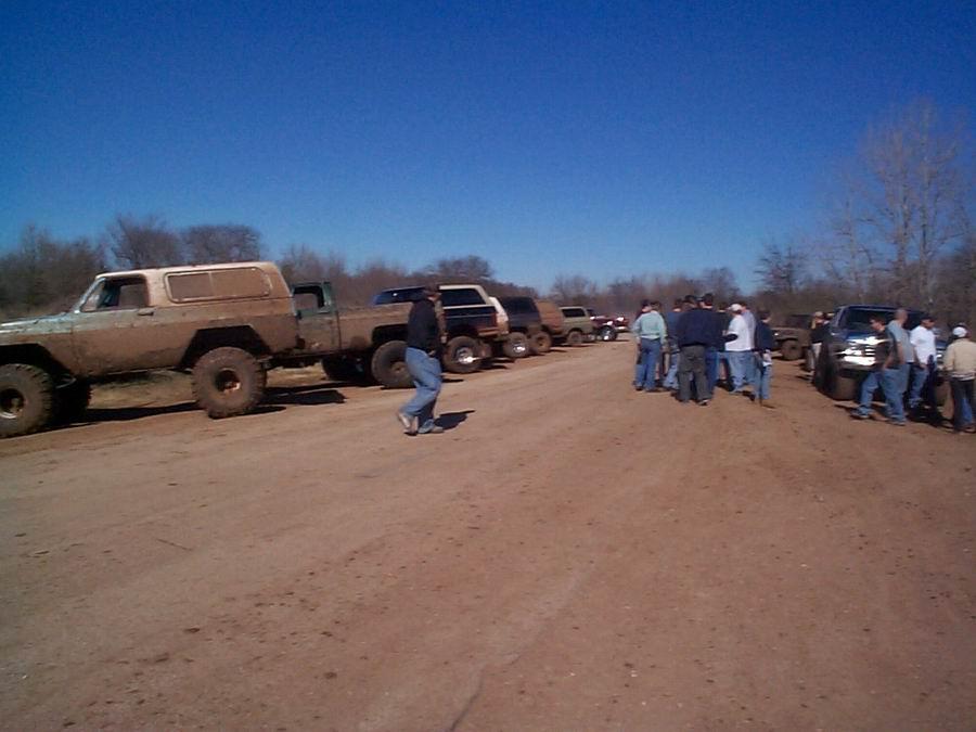Row of lifted mud trucks parked on a dirt staging area with people standing around