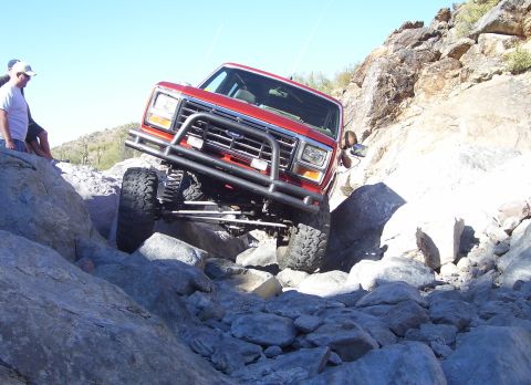 1984 Ford Bronco - A red off-road SUV tackles a challenging rocky terrain on a sunny day.