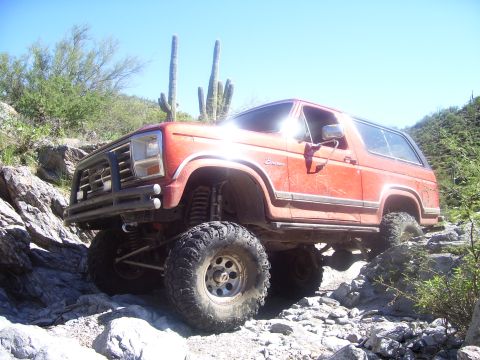 1984 Ford Bronco - This red SUV is navigating a challenging rocky trail under a clear blue sky.