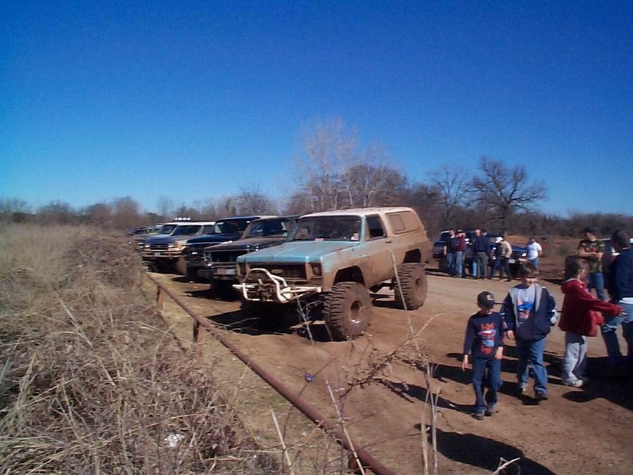 Pale blue lifted Chevy Blazer with brush guard parked among trucks as kids walk past