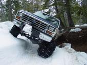 1986 Ford Bronco - This off-road vehicle is attempting to climb a snowy bank, showing its front grille and large tires.