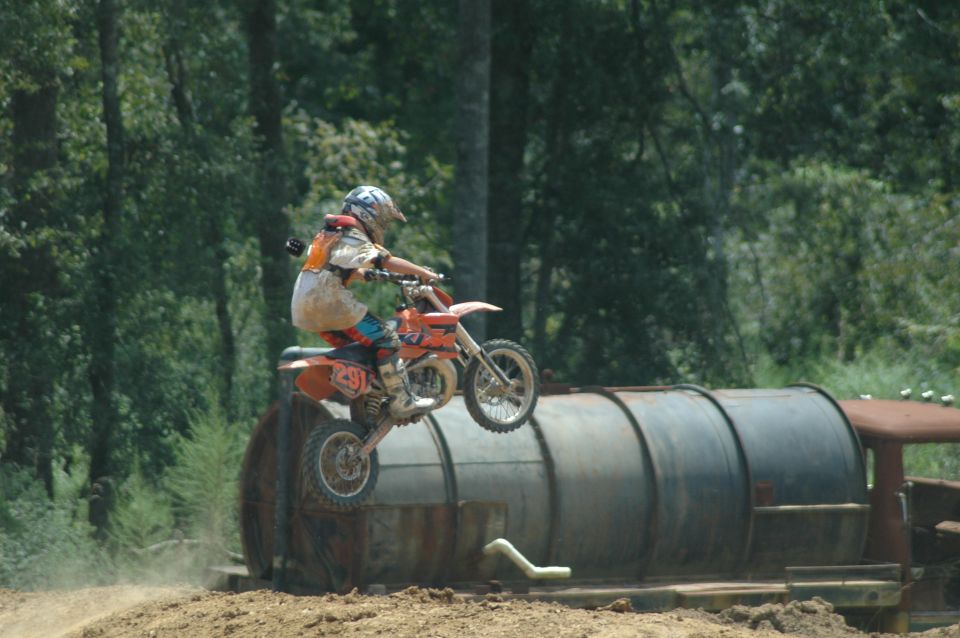 Rider on orange dirt bike #291 jumping over a rusty tank at a wooded motocross track