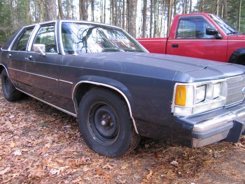 1991 Ford LTD Crown Victoria - Dark blue sedan parked in a wooded area next to a red pickup truck.