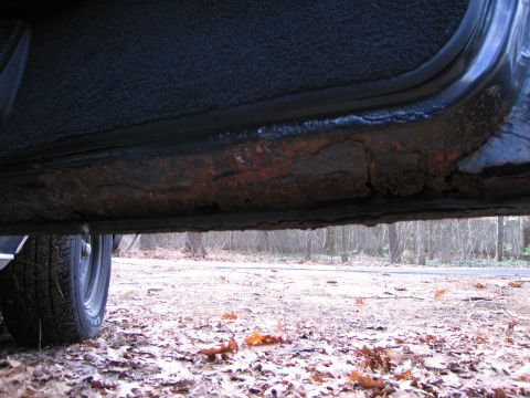 1991 Ford LTD Crown Victoria - The rocker panel of a vehicle shows significant rust and deterioration, with a tire visible on the left.
