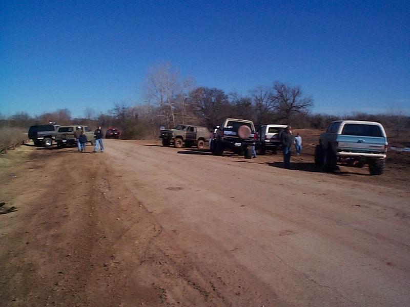 Line of off-road trucks and SUVs parked along a dirt road under clear blue sky with people milling around