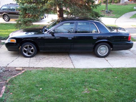 2001 Ford Crown Victoria - A black 2001 Ford Crown Victoria sedan is parked on a concrete driveway next to a lawn and a tree.
