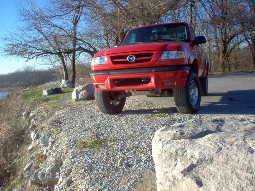 Red Mazda B3000 pickup truck parked on a gravelly area near some trees and water