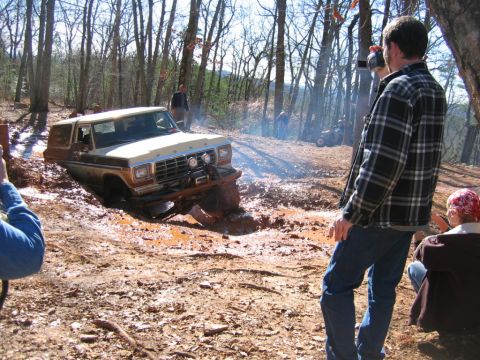 1992 Ford Mustang - A Ford Bronco is stuck in the mud while onlookers observe the scene in a wooded area.