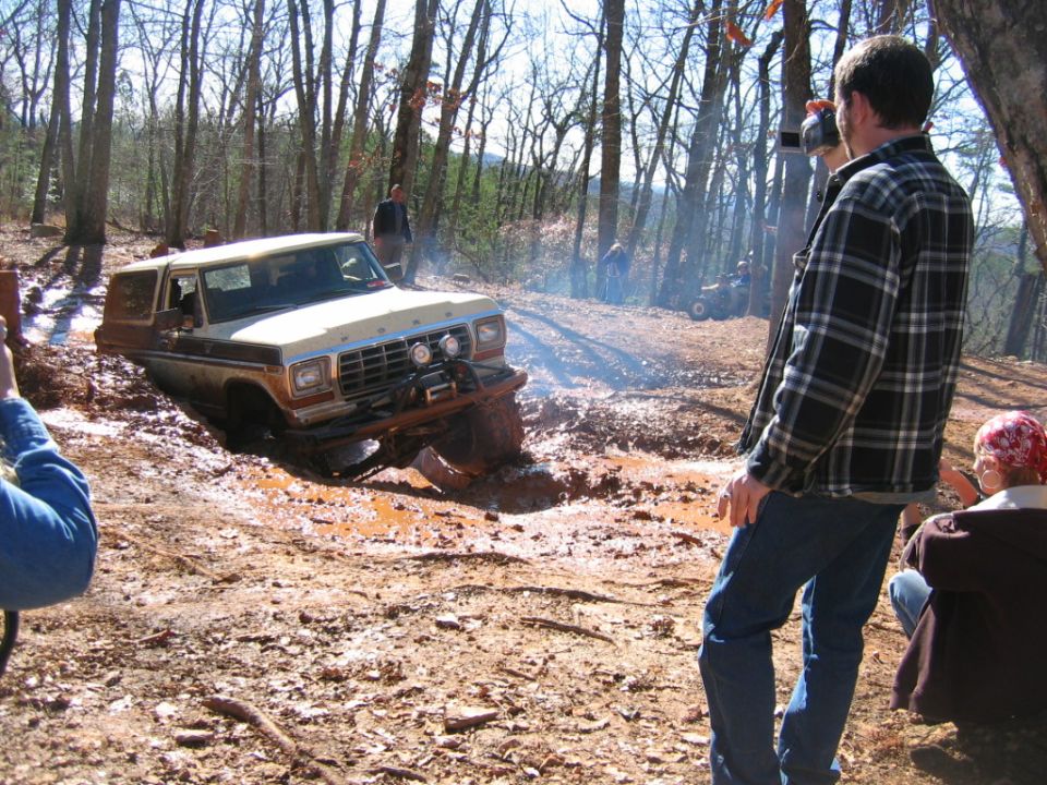 Tan and brown Ford Bronco stuck in mud, front end low, with people watching