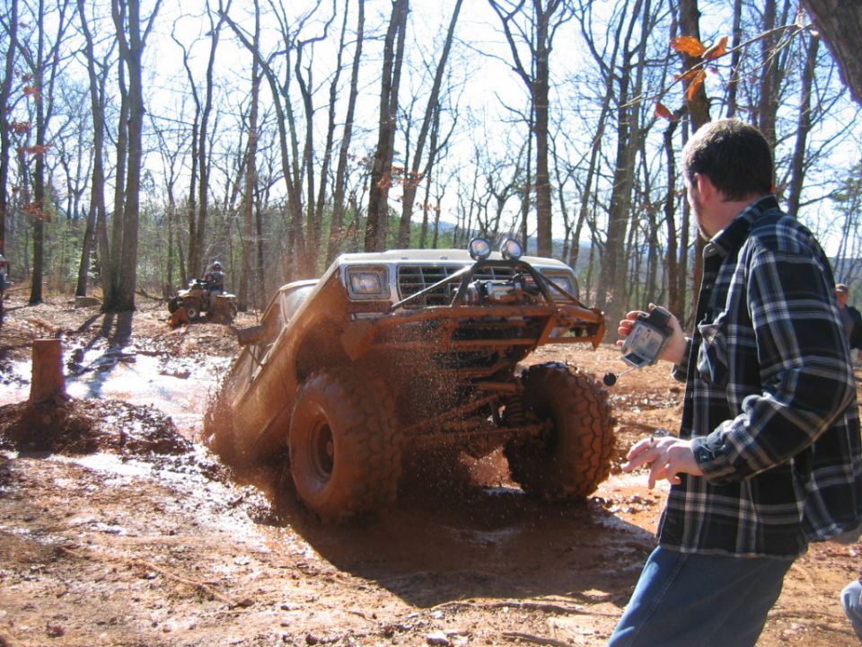 Off-road vehicle covered in mud, stuck in a muddy trail with trees in the background. Another ATV is visible in the distance