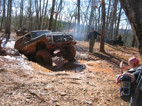 1992 Ford Mustang - An off-road SUV navigates a muddy trail in a wooded area, with other vehicles and spectators present.