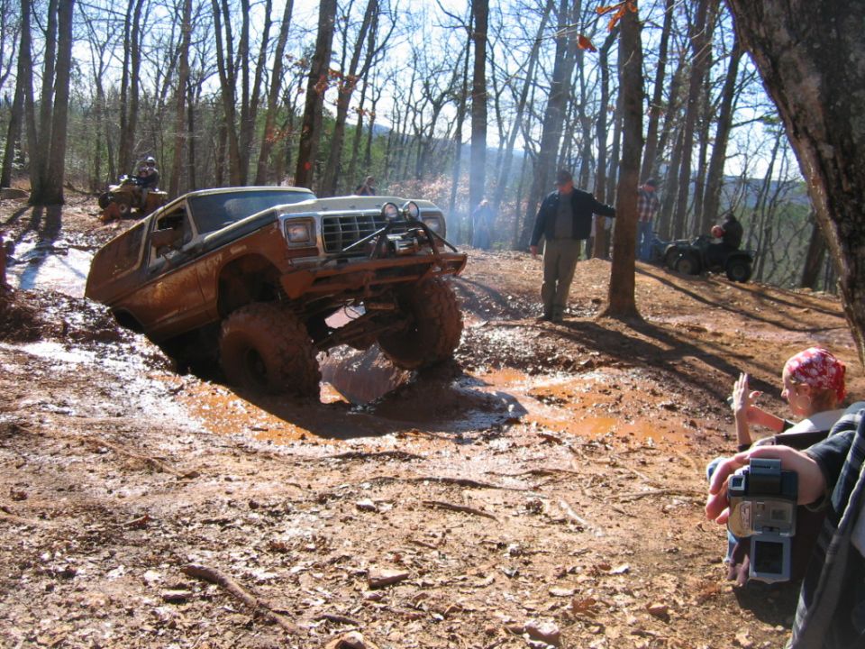 Off-road SUV with large tires stuck in mud on a wooded trail. Another ATV is visible in the background