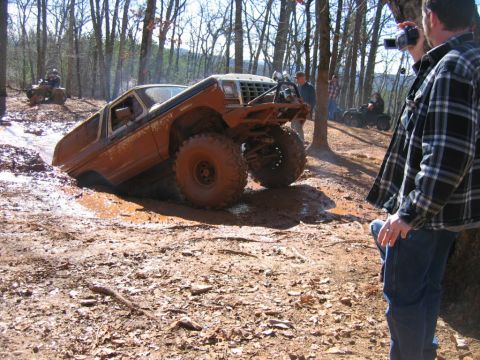 1992 Ford Mustang - An off-road truck is stuck in a muddy ditch while other ATVs and people watch in a wooded area.
